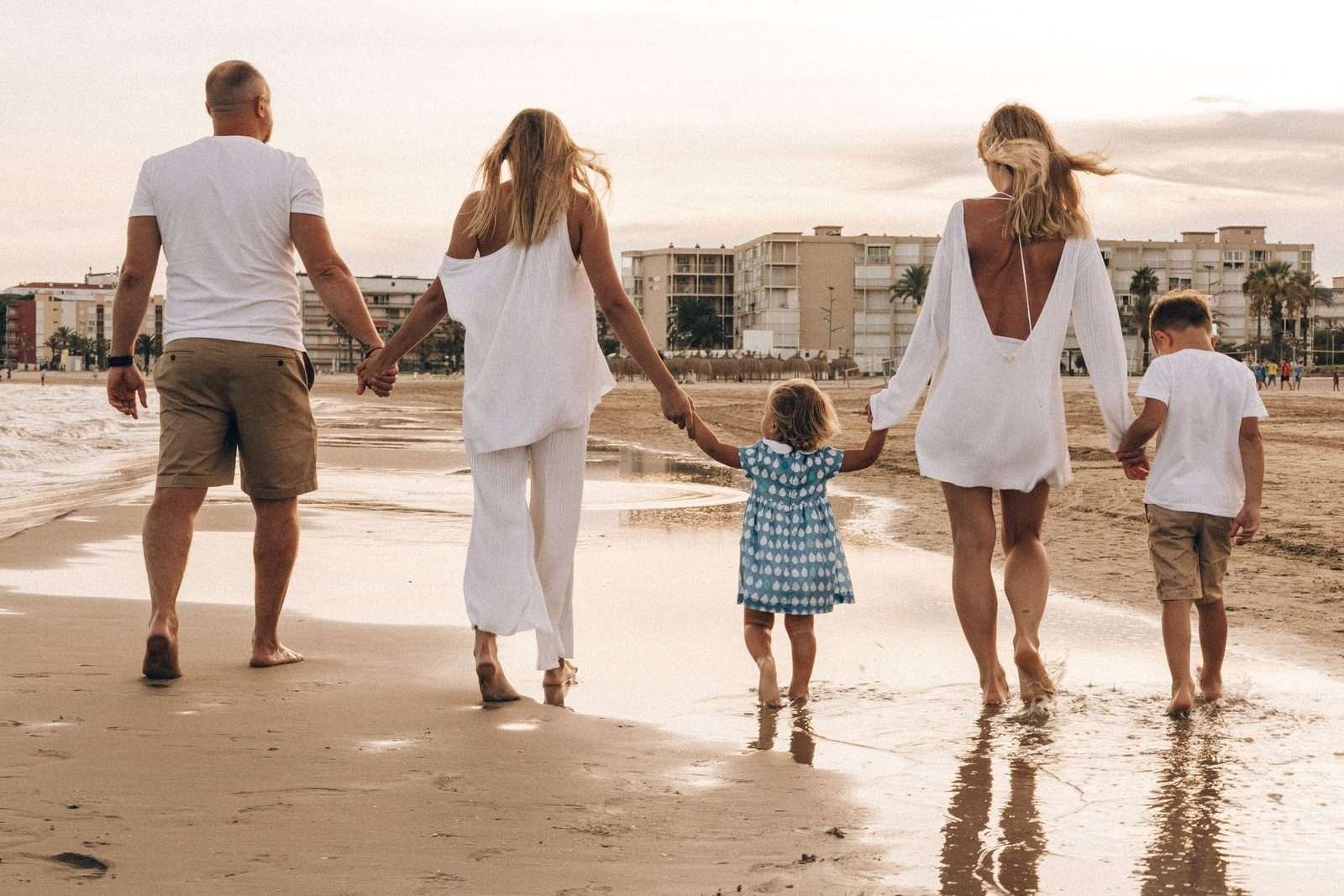 Família vestida de branco caminhando de mãos dadas pela beira da praia no fim de tarde